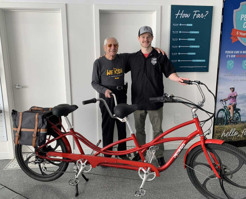 Two happy riders stand next to a Pedego tandem ebike. Indoor photo.