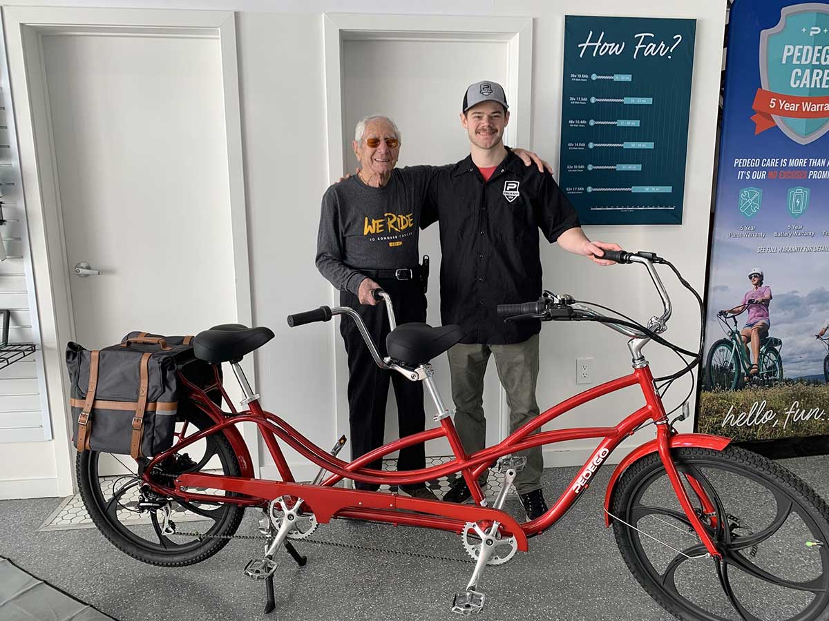 Two happy riders stand next to a Pedego tandem ebike. Indoor photo.