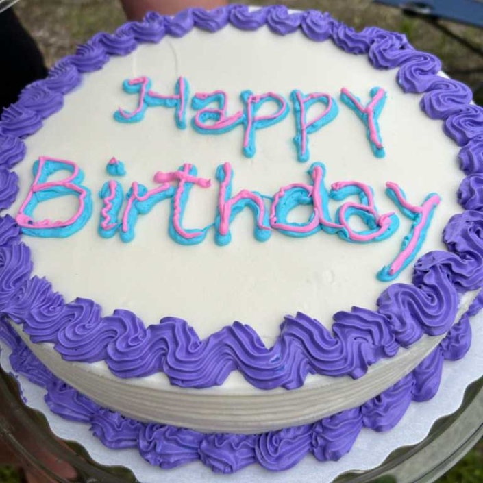 A Birthday Cake with Happy Birthday written on it. White with purple icing.