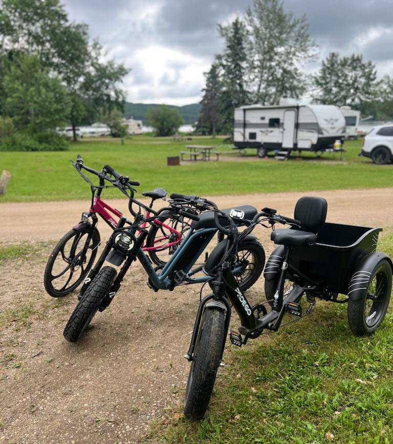 The Pedego Fat Tire Trike, Pedego Moto and Pedego Avenue are parked at a campground with an RV in the background.