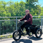 An 80-year-old man rides a Pedego Fat Tire Trike on a paved path by a railing.