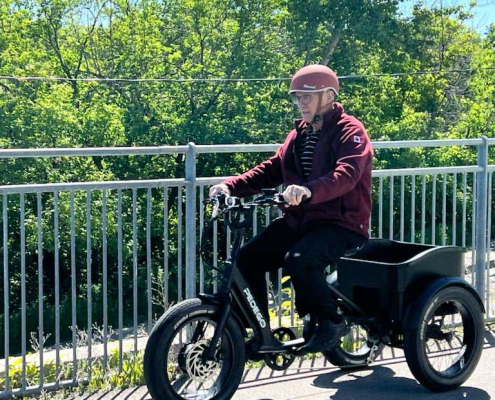 An 80-year-old man rides a Pedego Fat Tire Trike on a paved path by a railing.